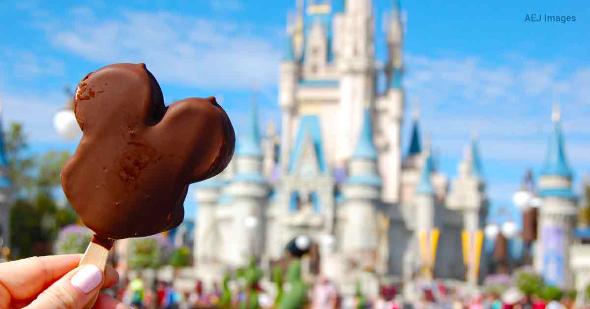A hand with white nail polish holds a complete Mickey ice cream bar, left of center. In the background, you can see a blurred Cinderella's castle and talking patrons