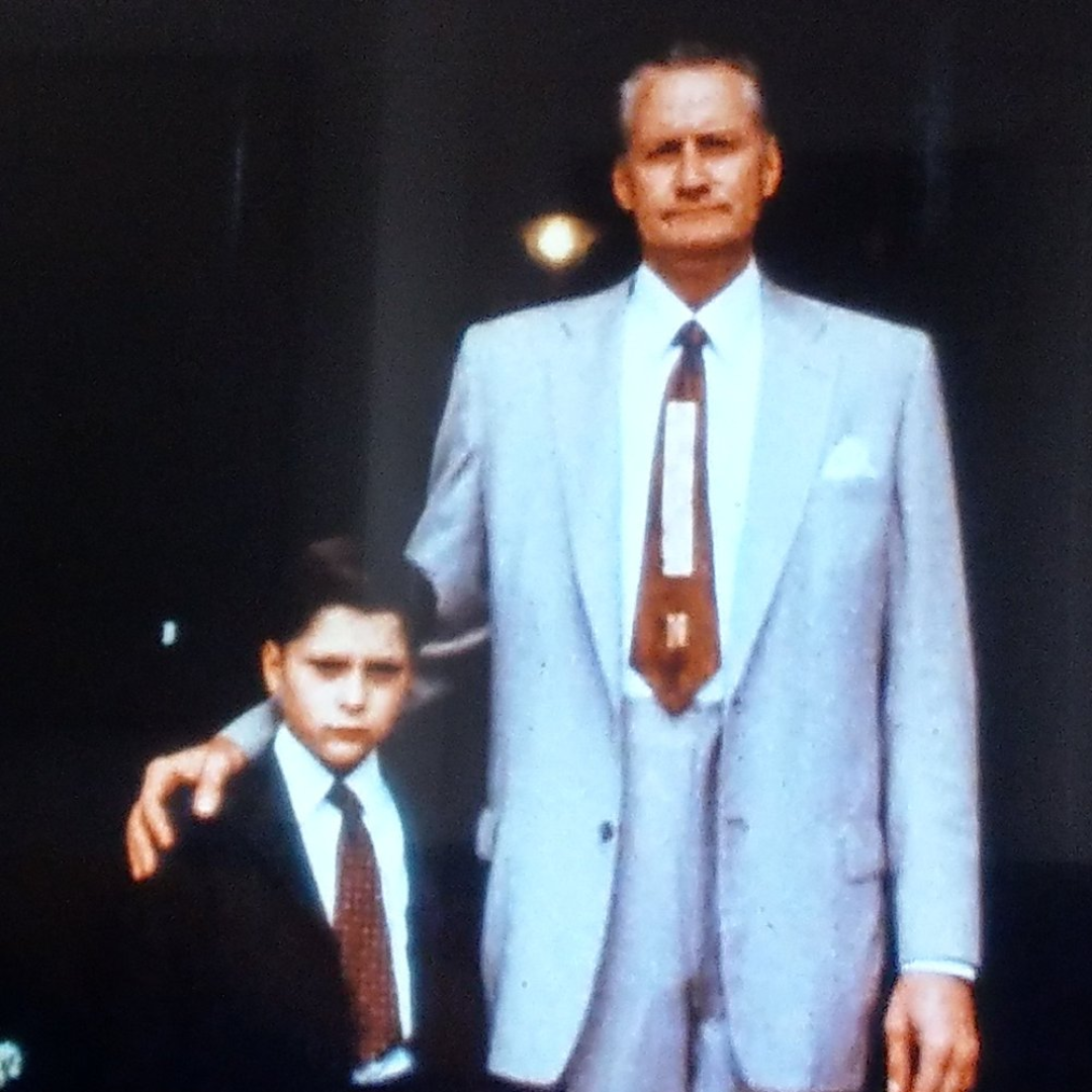 Charles Martinet in a blue/gray suit with a red tie with his hand on a young boy's shoulder who is also wearing a suit.