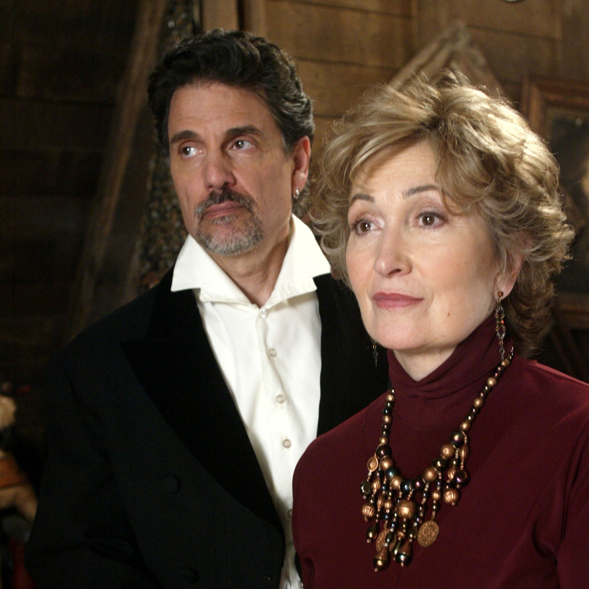 Chris Sarandon as the Necromance. He is wearing a white button up and a black jacket. He is standing next to a woman in a maroon turtle neck with a bulky brass necklace.