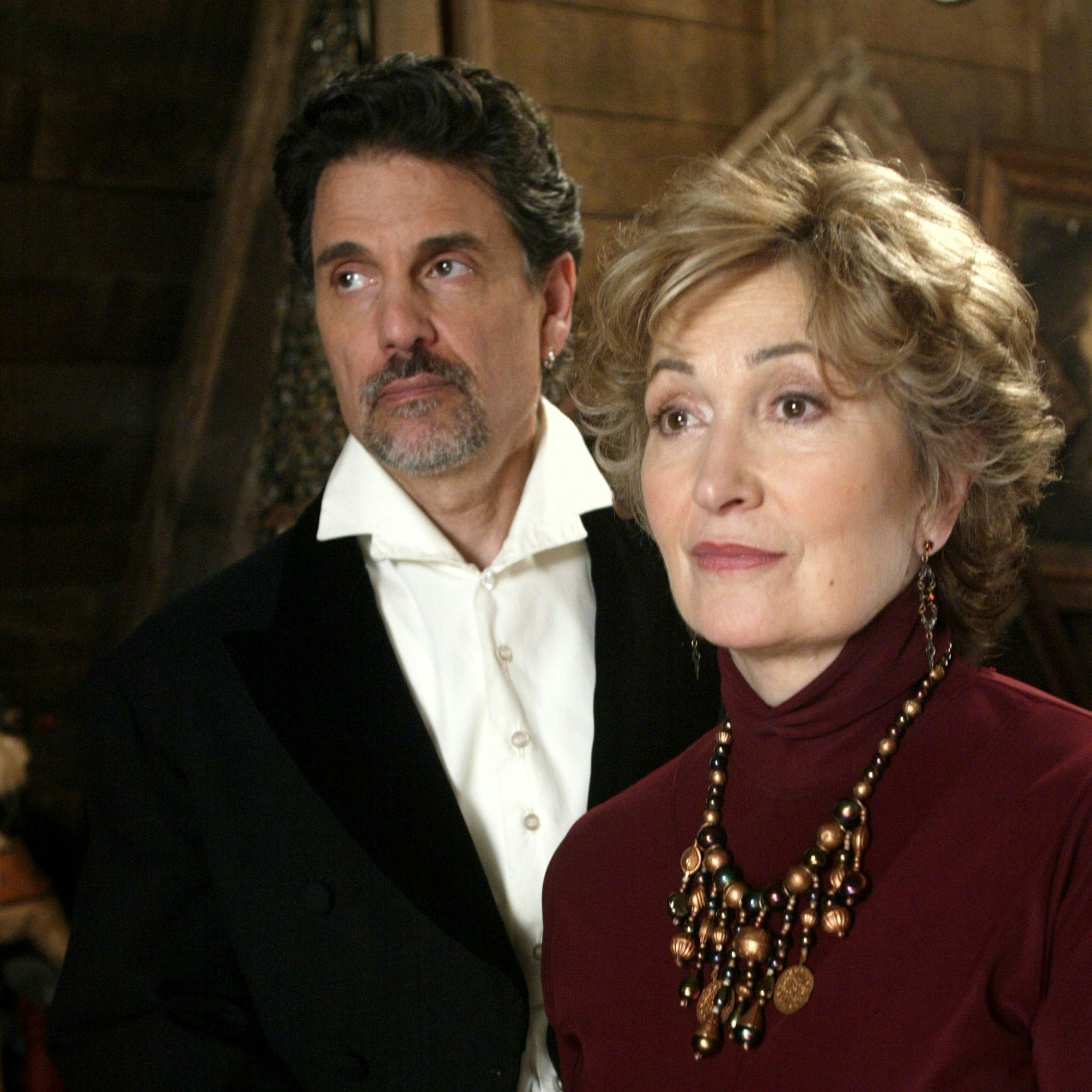 Chris Sarandon as the Necromance. He is wearing a white button up and a black jacket. He is standing next to a woman in a maroon turtle neck with a bulky brass necklace.
