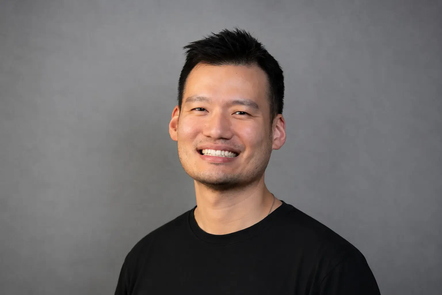 Foto de perfil em formato circular de homem asiático sorrindo, com cabelo curto escuro, vestindo camiseta preta contra fundo cinza neutro.