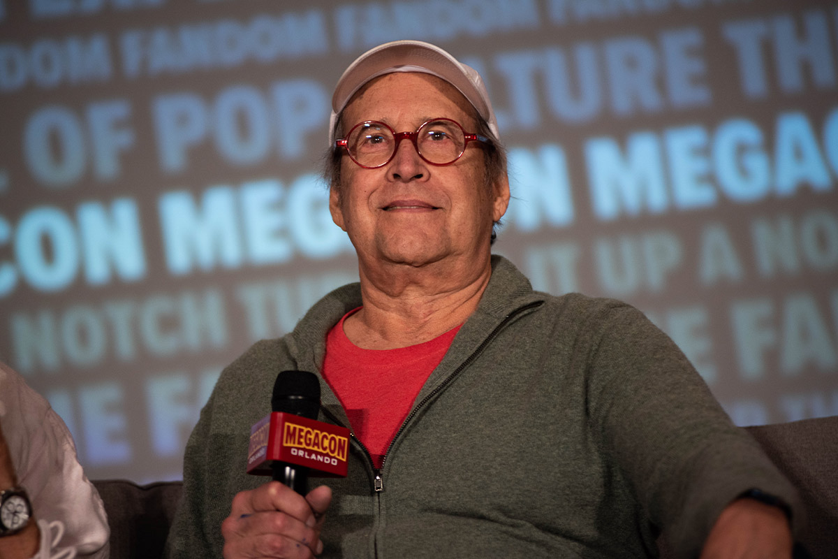 Chevy Chase smirks while sitting on stage in a green sweater