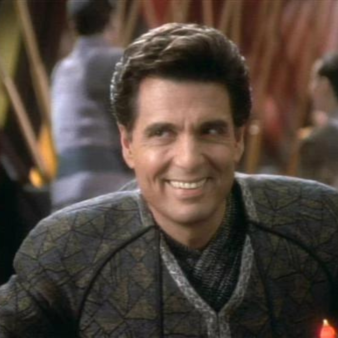Chris Sarandon is smiling. He is wearing a dark geometric shirt.