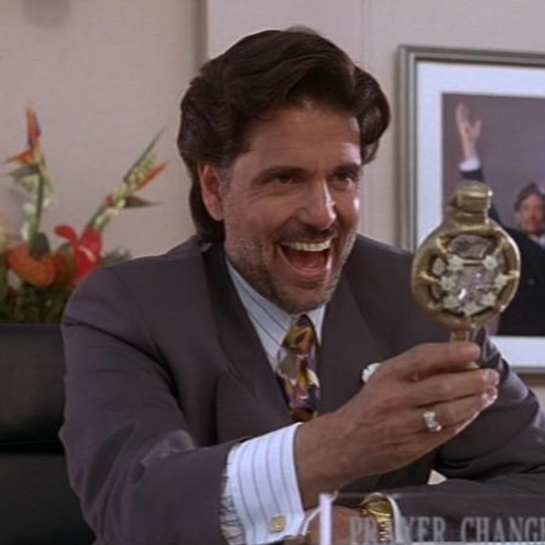 Chris Sarandon holding this metal thing. He is smiling and laughing sinisterly. He is wearing a brown suit. He is in an office with a flower arrangement and a picture of himself behind him.
