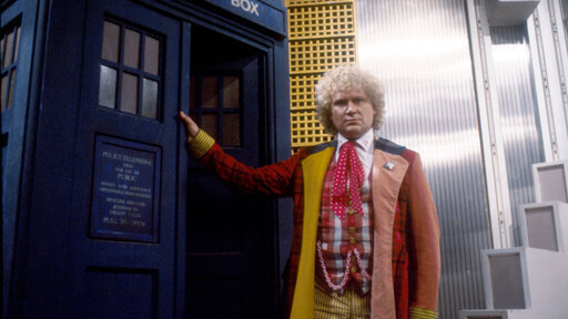 The Doctor stands in the doorway to the TARDIS, his face is stern while his clothing boisterous and colourful.  