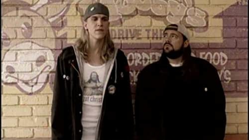 Jay and Silent Bob standing in front of a Mooby's sign painted on a brick wall. There is a cartoonish cow to the left of the pair. Silent Bob is wearing dark clothing and his hat is on backward. Silent Bob is looking up at Jay, who is wearing layers of jackets and shirts on top of a white tee with a image on it.