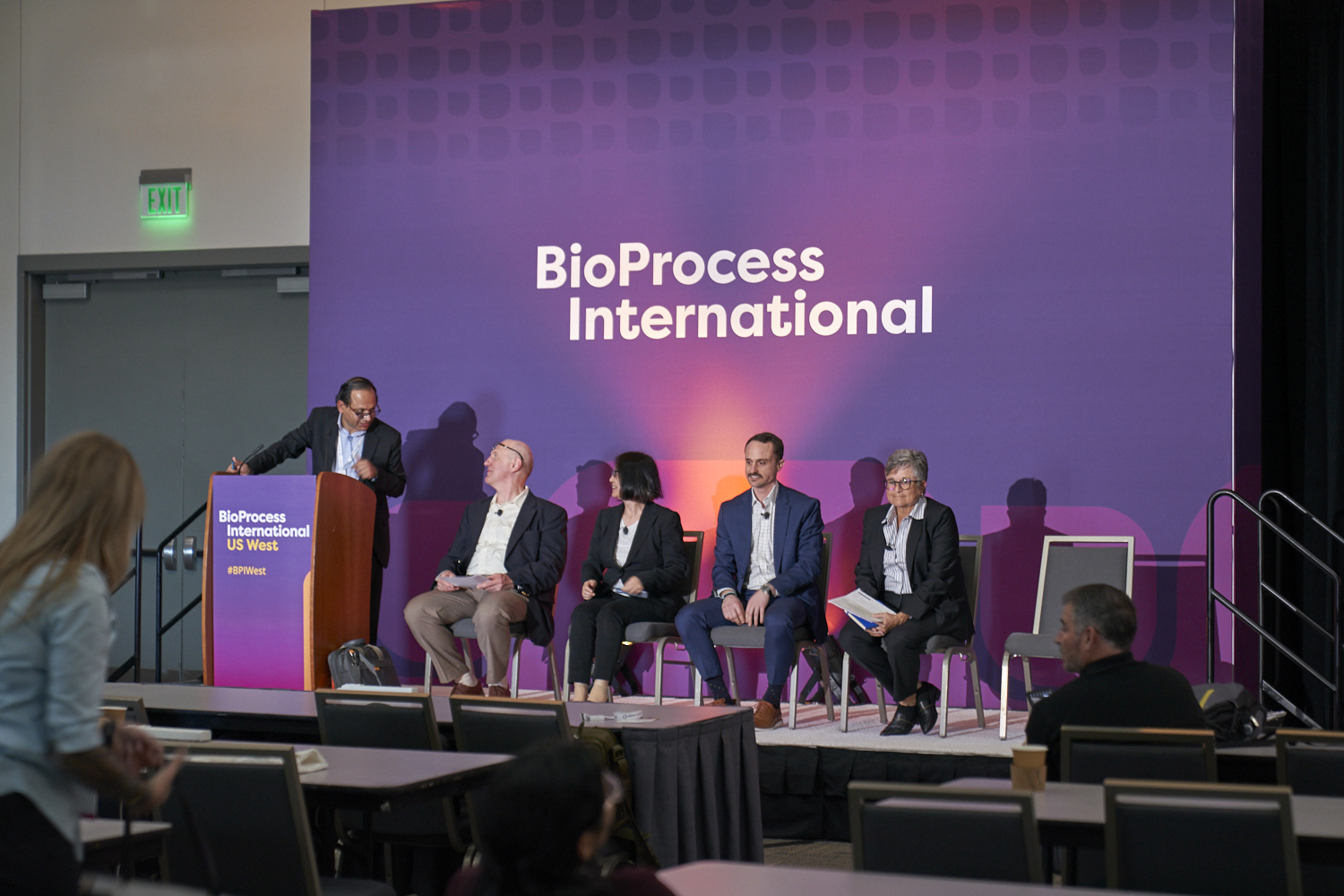4 speakers sit on stage and are asked questions by a host standing at a podium at BioProcess International