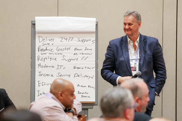 Photograph of an IT support leader in a professional business suit conducting an interactive session for conference attendees, standing next to a presentation board featuring trending IT service management topics, illustrating peer-to-peer knowledge sharing at HDI Service & Support World.