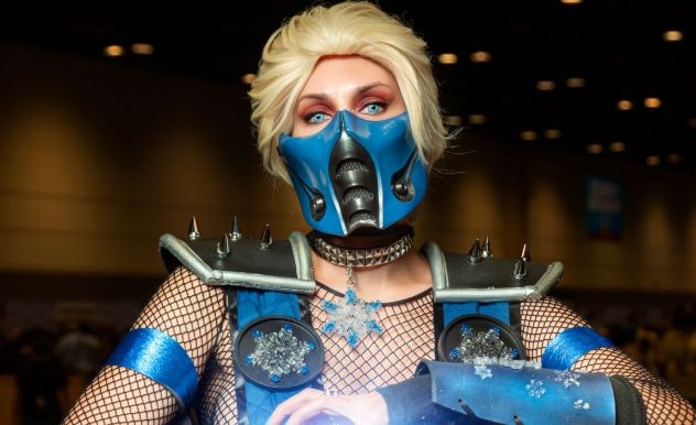 A cosplayer in a blonde with with a hard plastic blue and black mask, blue straps with snowflakes on them and a snowflake necklace