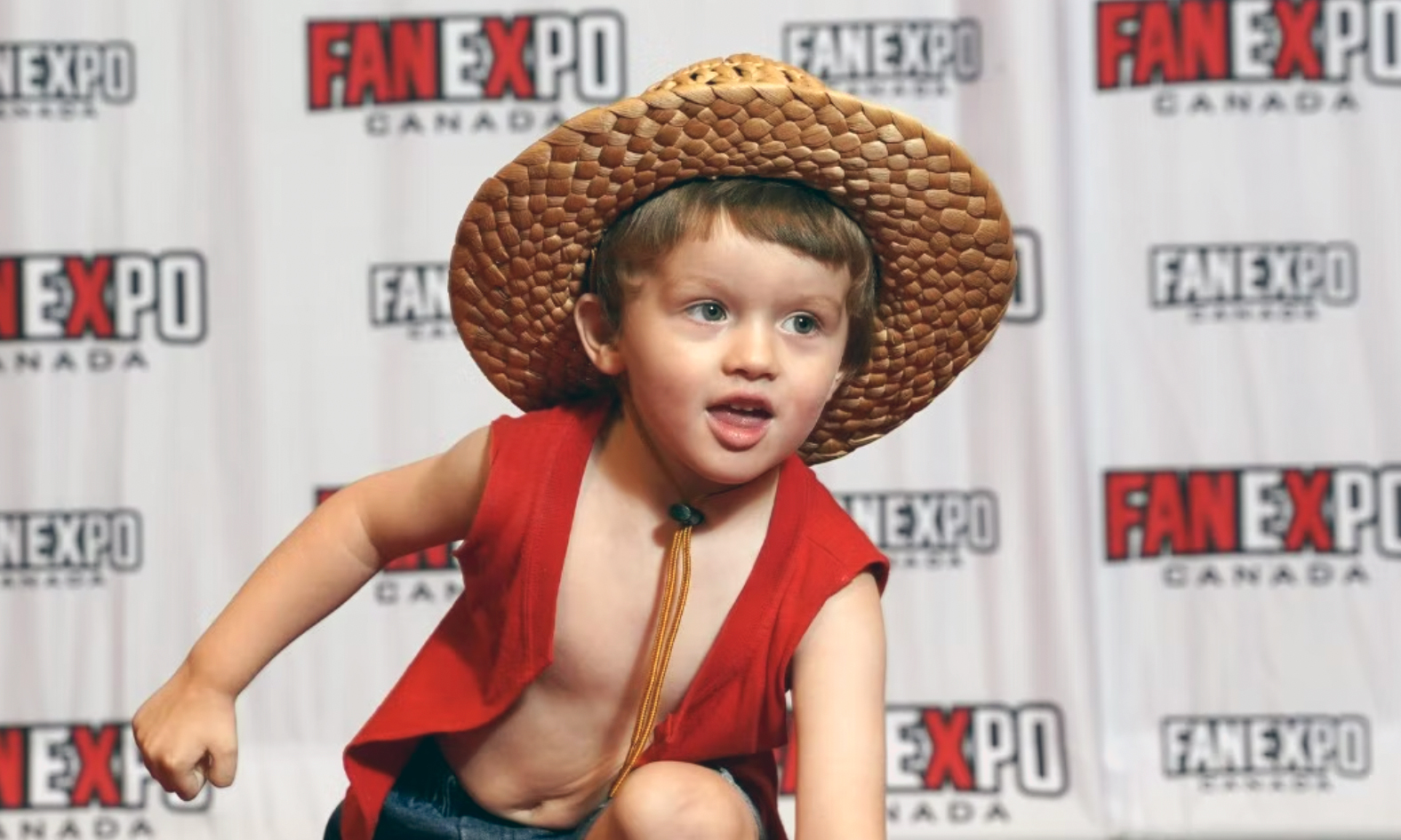 A young boy poses on the red carpet in a red vest with blue jean shorts and a brown straw hat.