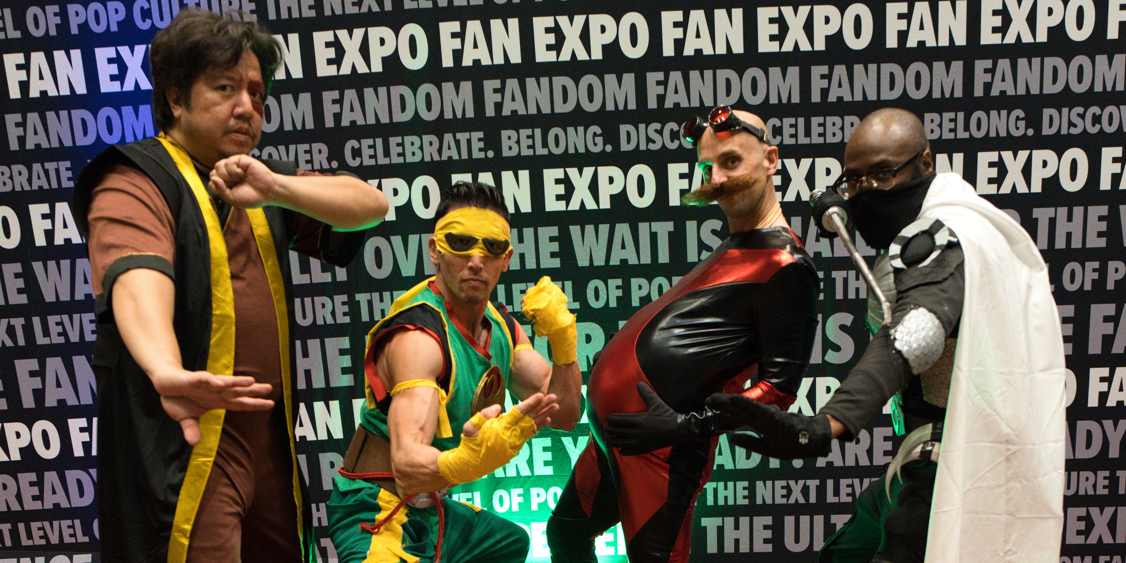A cosplayer in a fantasy-inspired costume with long lavender pigtails and large elf ears speaks into a microphone while leading a panel at FAN EXPO. Seated behind a table with a laptop, they address a room of attendees, one of whom has their hand raised to ask a question. The backdrop features repeating FAN EXPO and “For the Fans” logos.