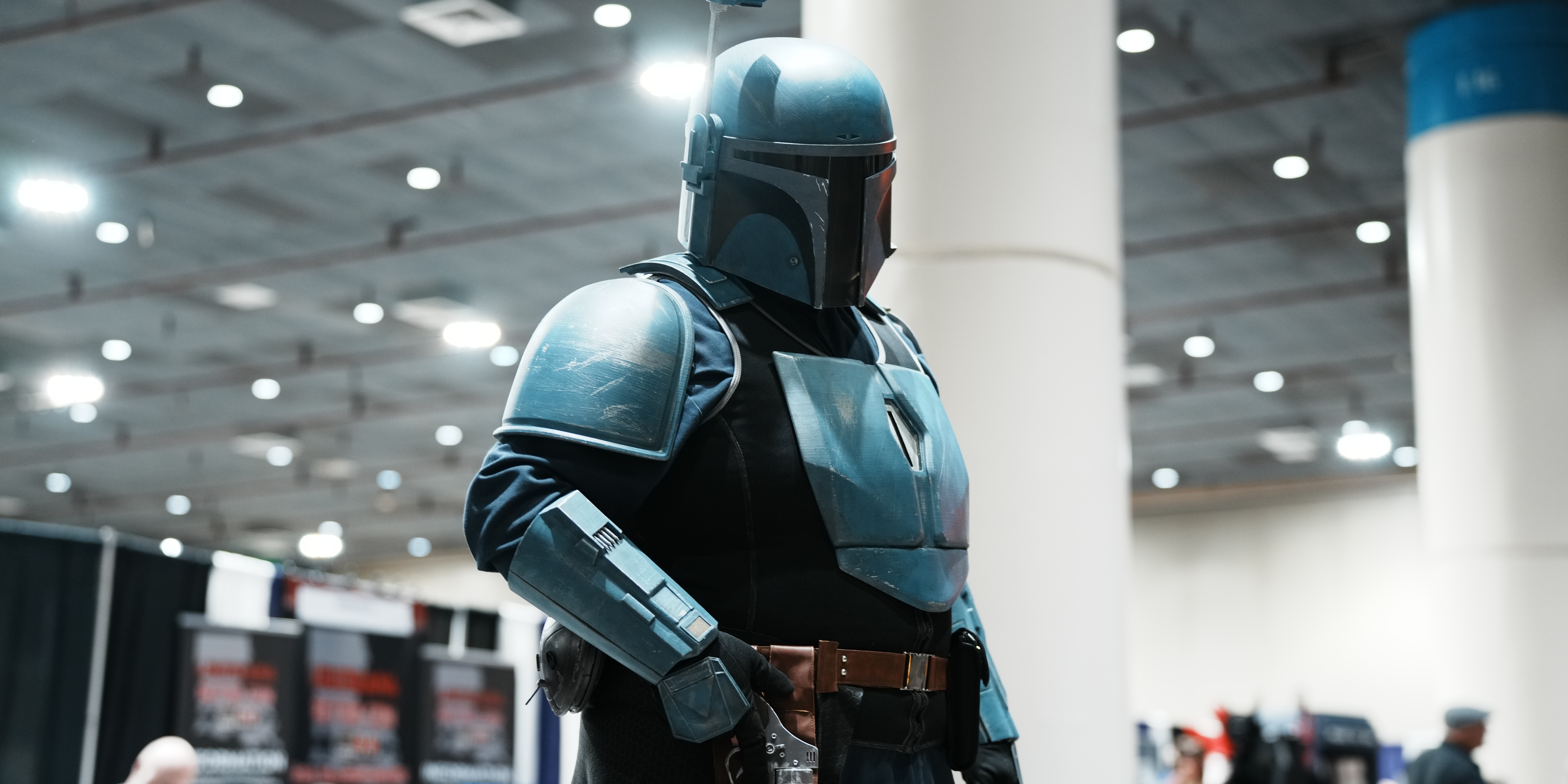 A cosplayer in a fantasy-inspired costume with long lavender pigtails and large elf ears speaks into a microphone while leading a panel at FAN EXPO. Seated behind a table with a laptop, they address a room of attendees, one of whom has their hand raised to ask a question. The backdrop features repeating FAN EXPO and “For the Fans” logos.