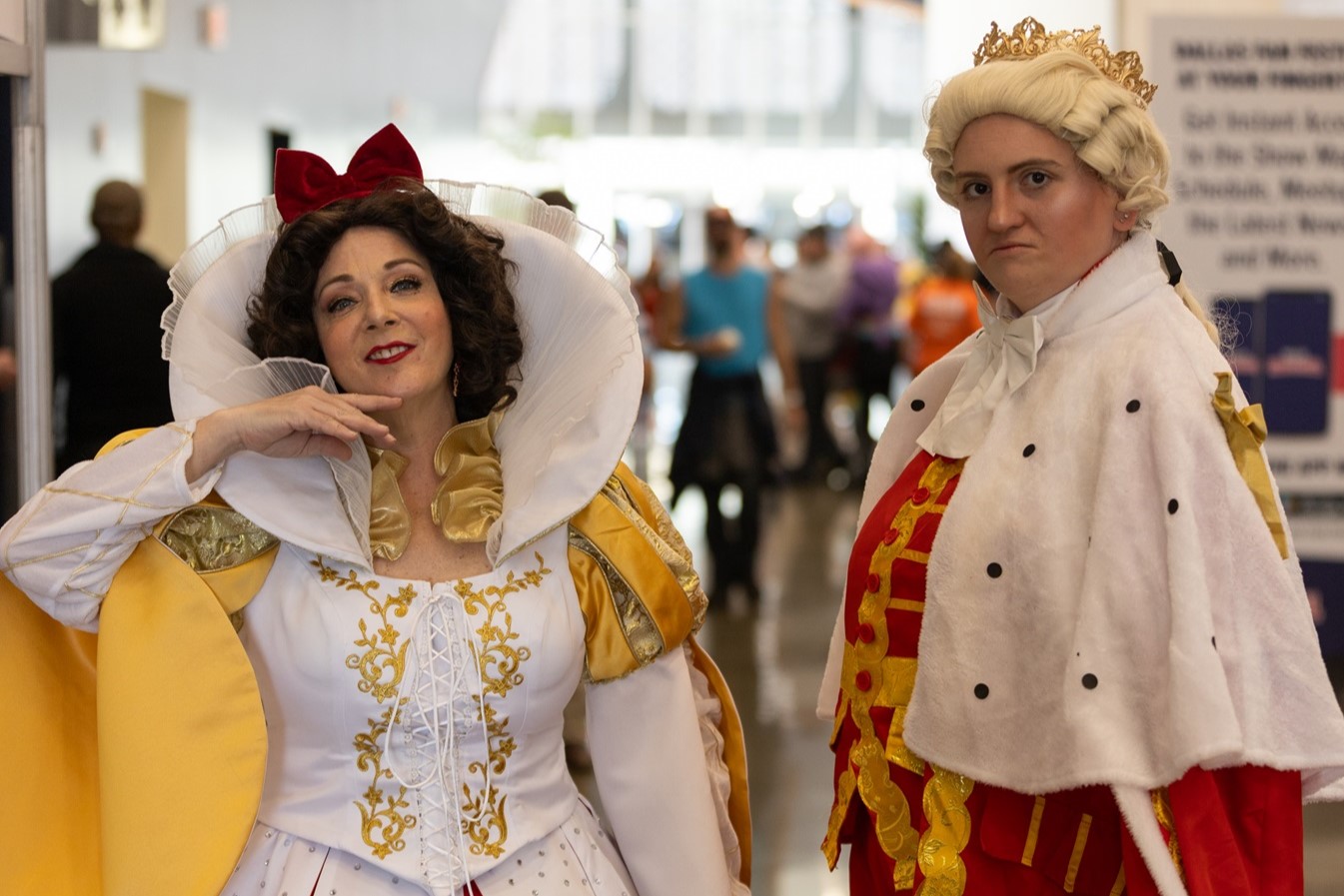Two cosplayers inside the expo hall dressed as Snow White and King George III, with Snow White smiling playfully and King George giving a serious expression.