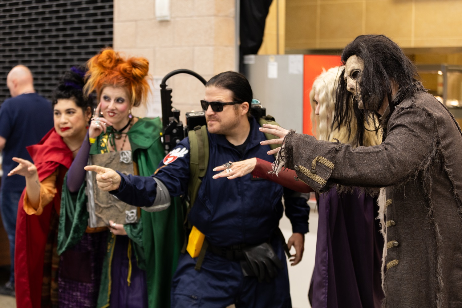 A group of cosplayers dressed as the Sanderson Sisters from Hocus Pocus, joined by a Ghostbuster and other spooky characters, playfully posing in character inside the expo hall.