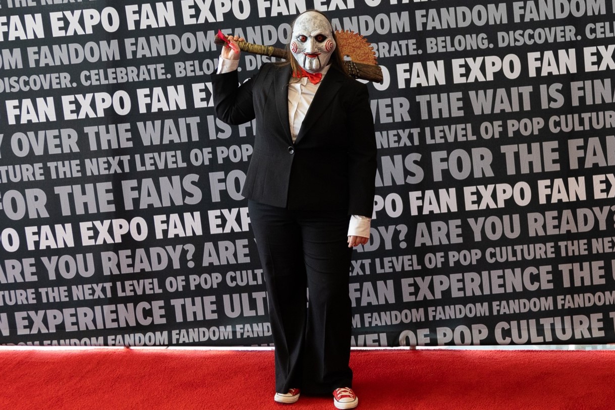 A cosplayer dressed as the Jigsaw puppet from Saw, wearing a suit and mask while holding a large prop saw, posing menacingly against the FAN EXPO backdrop.