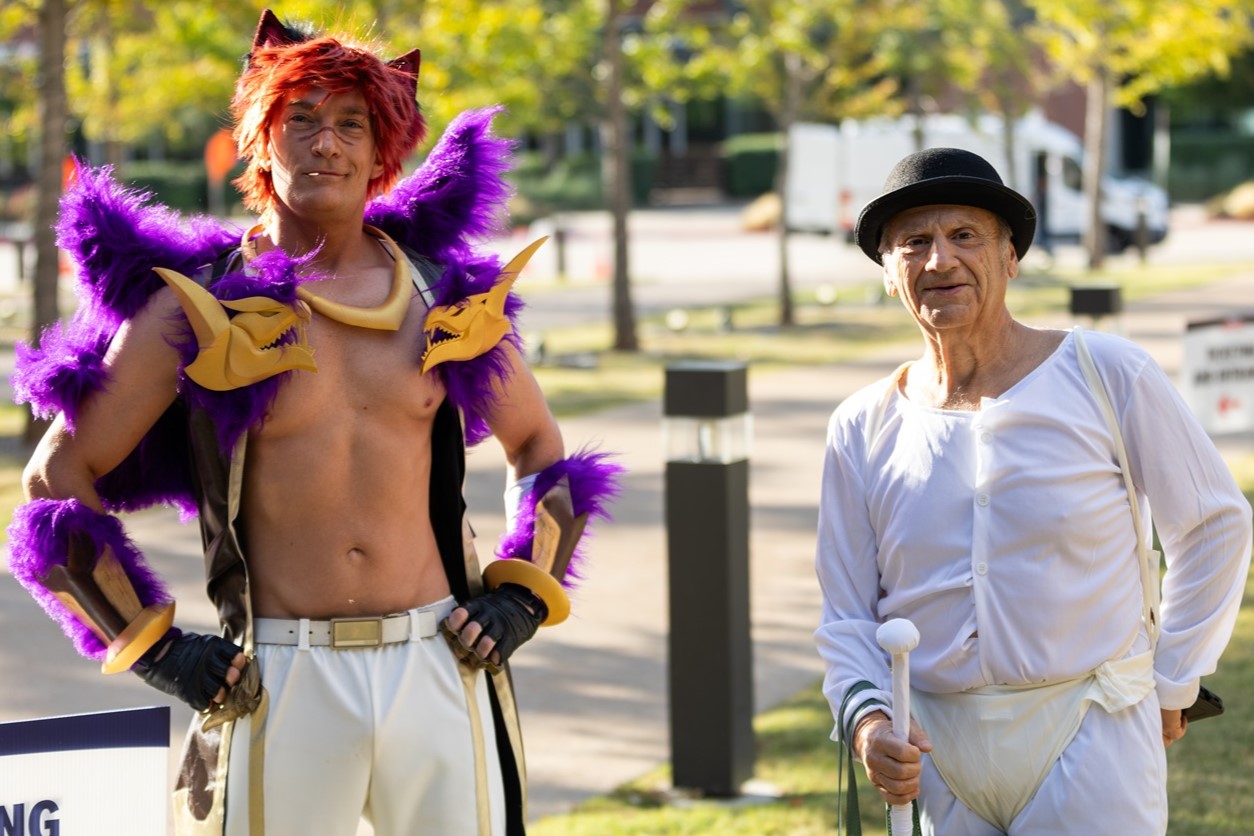 Two cosplayers posing outdoors. One is dressed in a purple and yellow fantasy-inspired costume with fur accents and horns, while the other is wearing a bowler hat and white attire resembling a character from A Clockwork Orange.