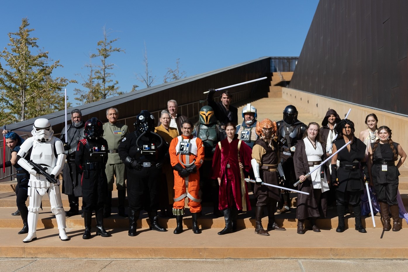 A large group of Star Wars cosplayers on an outdoor staircase, including characters like Stormtroopers, Rebel pilots, Mandalorians, and Jedi, standing in costume under a bright blue sky.