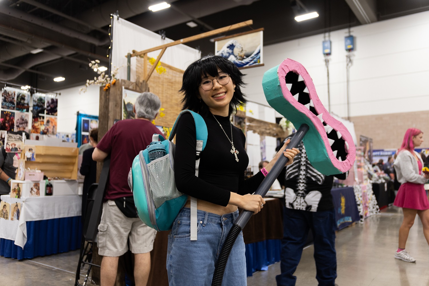 A young cosplayer inside the expo hall holding a large pink and green prop weapon, with a playful smile and a turquoise backpack.