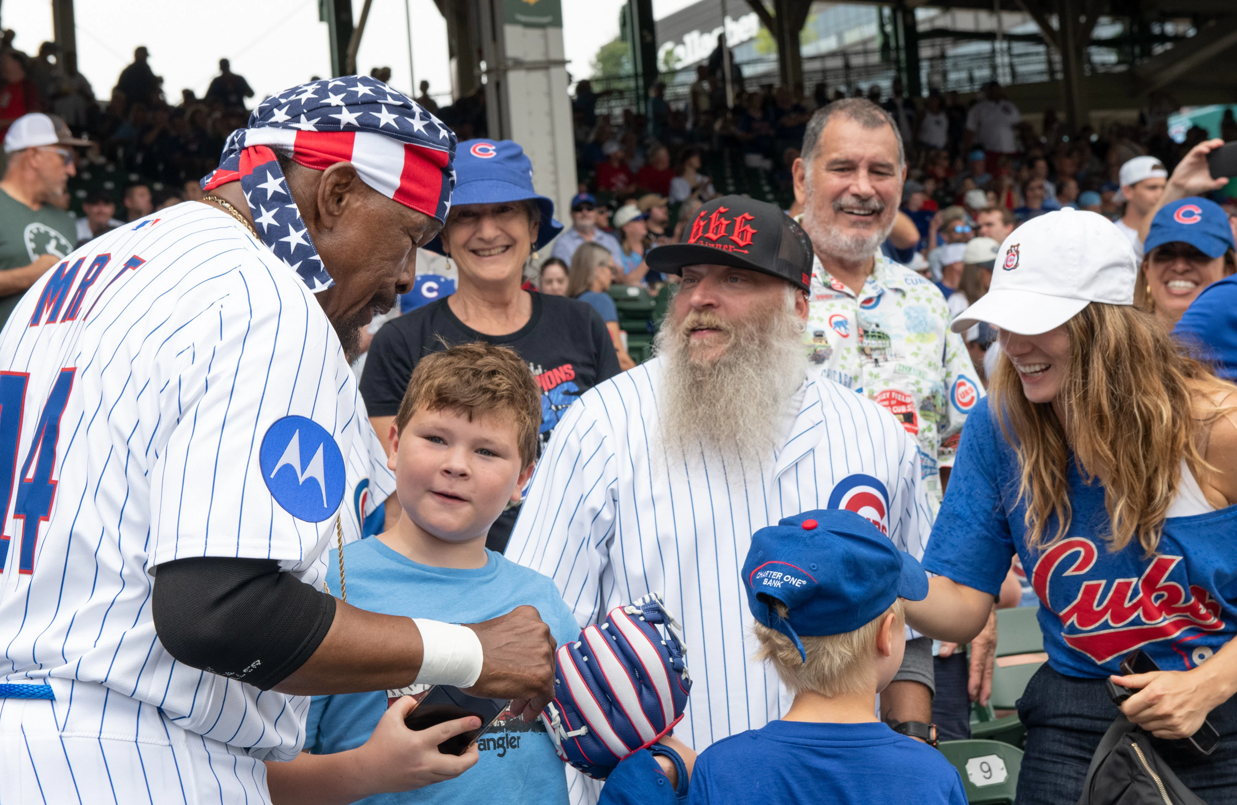 Hayden Christensen shakes hands with Clark the Cub mascot near the field, both wearing Cubs jerseys as the stadium crowd looks on.