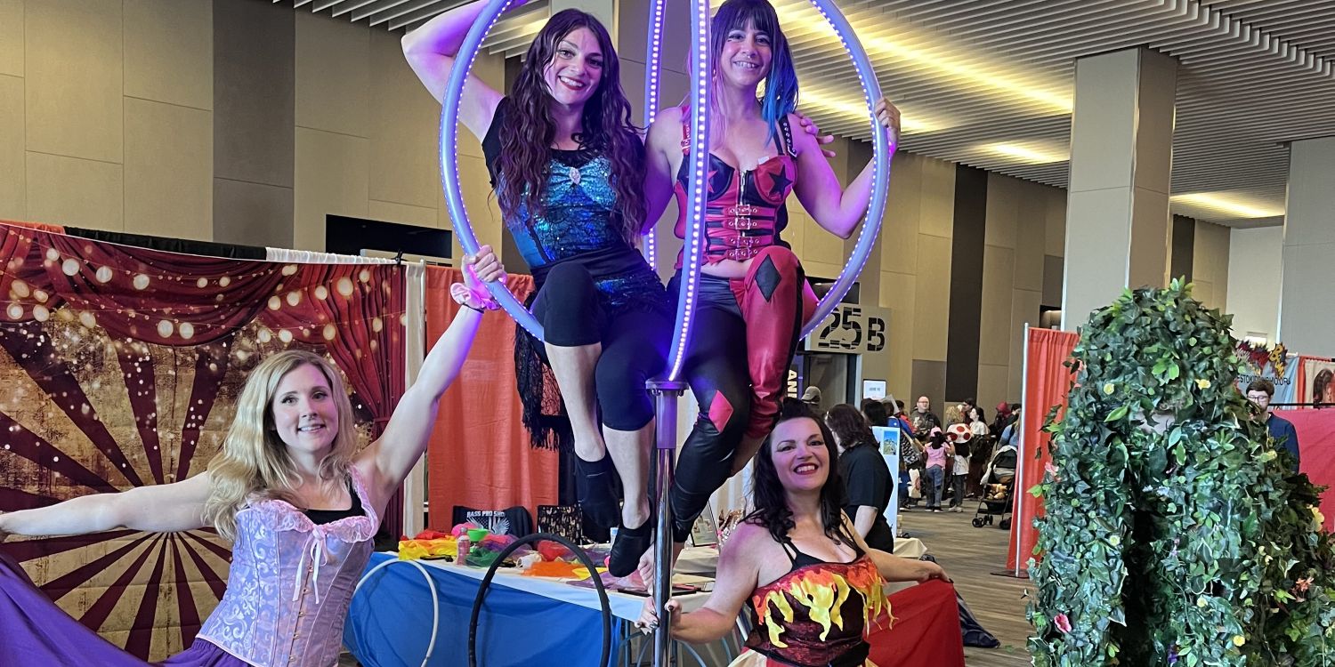 Four women dressed in colorful costumes pose in a lively convention setting. Two women sit on a glowing aerial hoop, while another spreads a flowing purple ribbon. A person dressed as a walking bush adds whimsy to the scene.