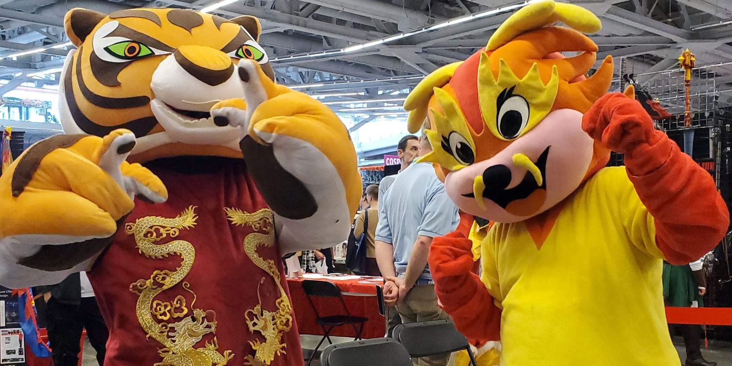 Two mascots, one dressed as a tiger in a red and gold embroidered vest and another as a bright yellow and orange fox-like character, strike playful poses at a convention with vendor booths in the background.