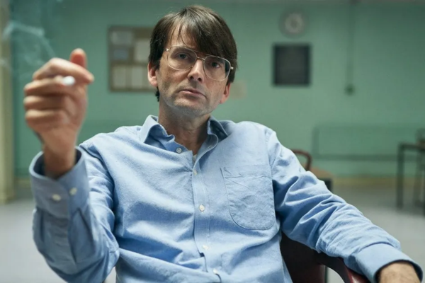 In a blue wrinkled button up shirt, David Tennant is smoking a cigarette and sitting in a green room. His hair is brushed downward and is wearing wired 70's glasses