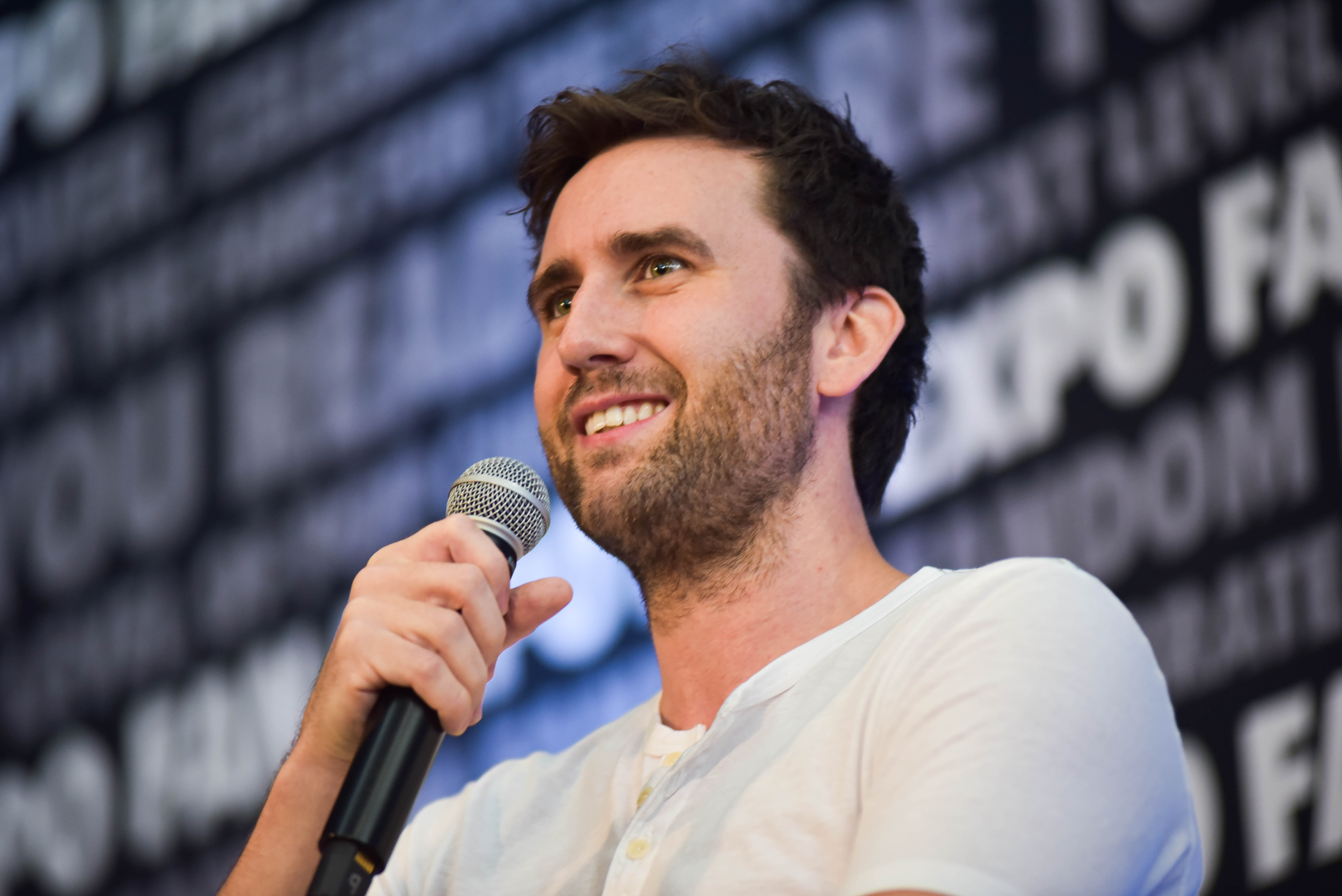 Matthew Lewis smiling as he holds his microphone close to his face at his panel.
