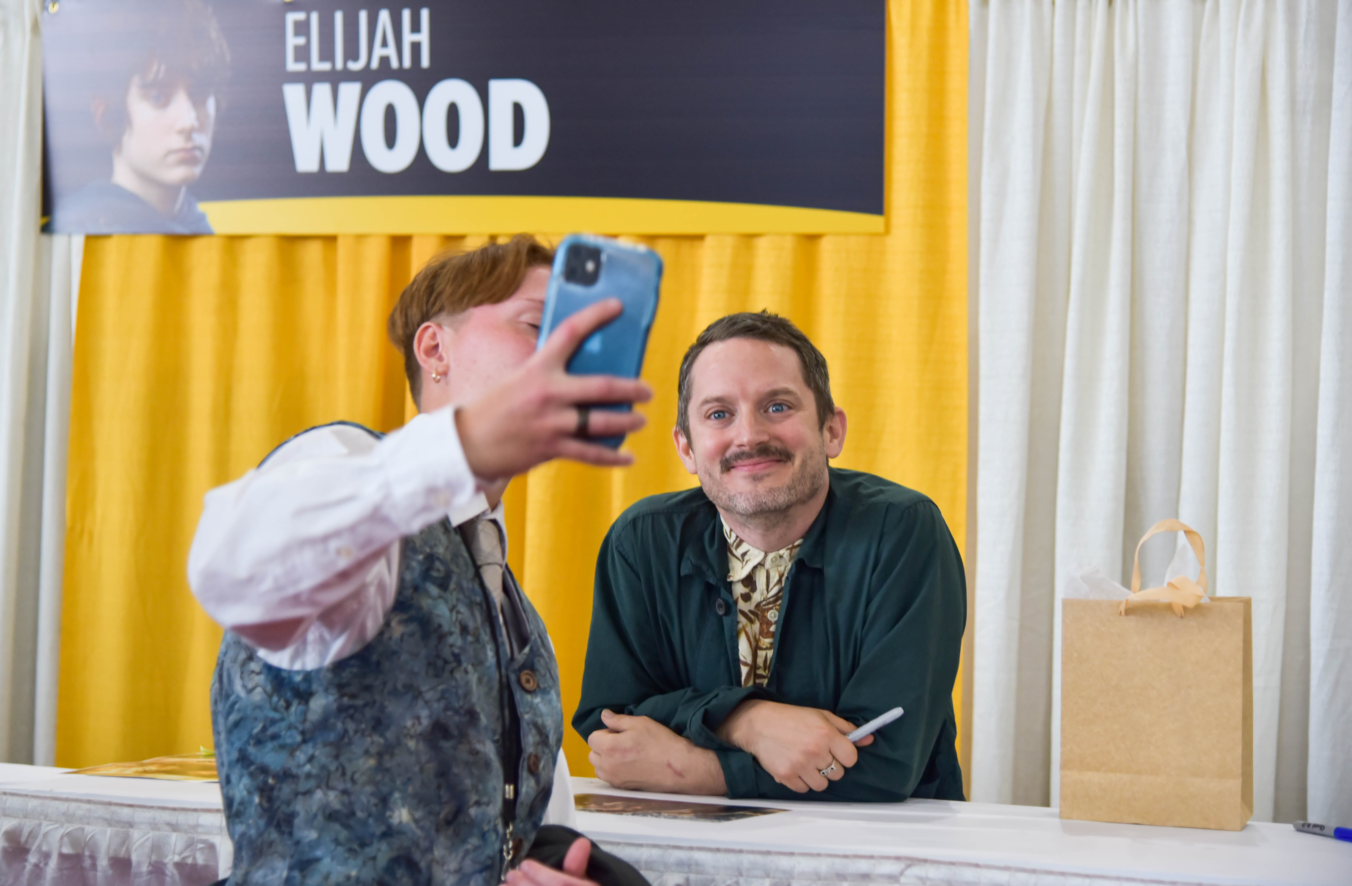 Elijah Wood posing at his table, all while a fan takes a selfie of them both.