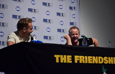 Dominic Monaghan and Billy Boyd sitting at a black table, looking at each other and laughing