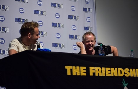 Dominic Monaghan and Billy Boyd sitting at a black table, looking at each other and laughing