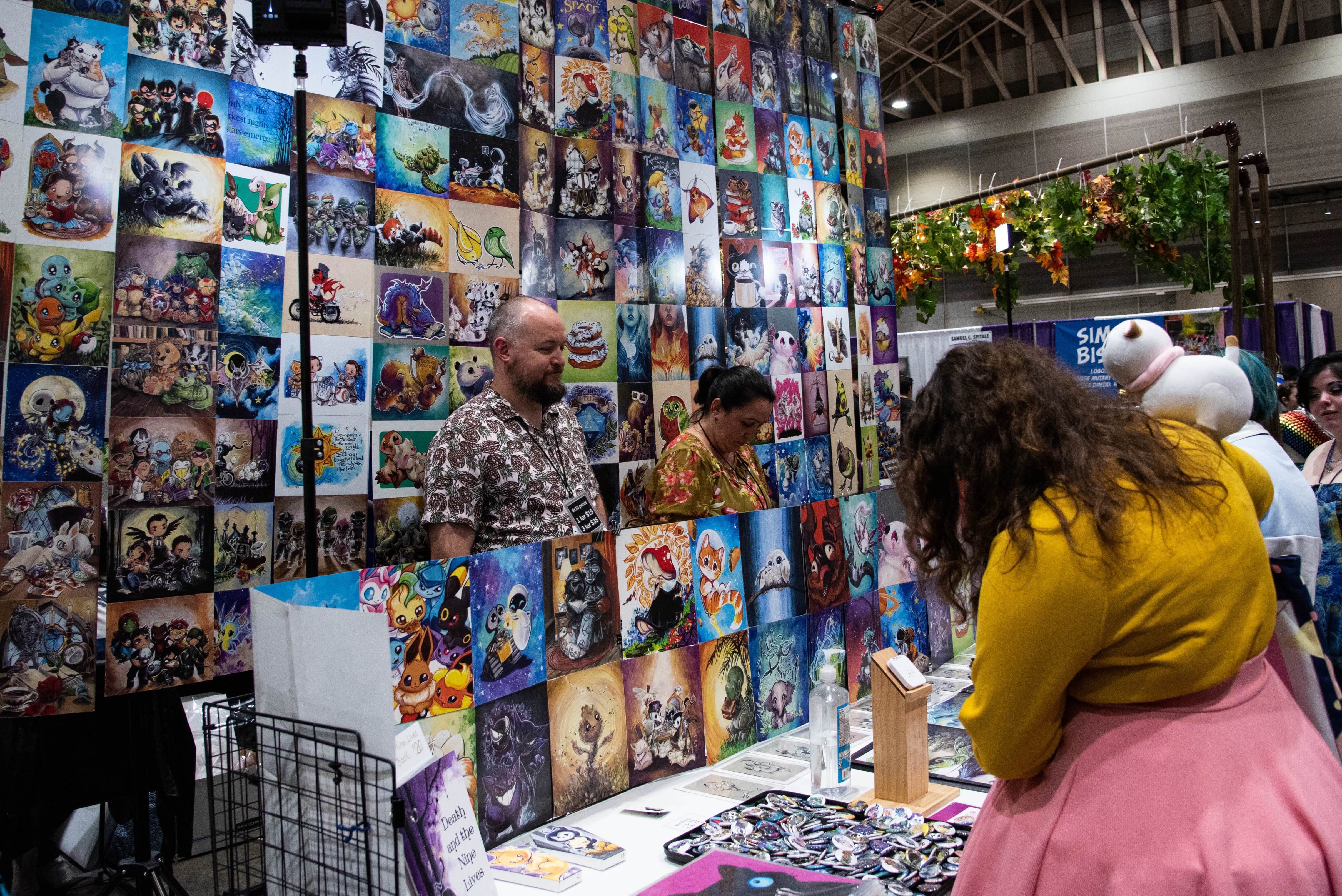 A Bee and PuppyCat cosplayer shopping for prints at an artist alley booth.