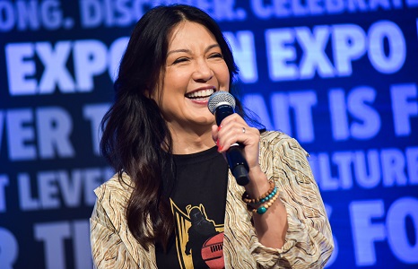 Ming Na-Wen holding a microphone and laughing. Wearing a black t-shirt with the Mandalorian on it, with a beige cardigan over it.
