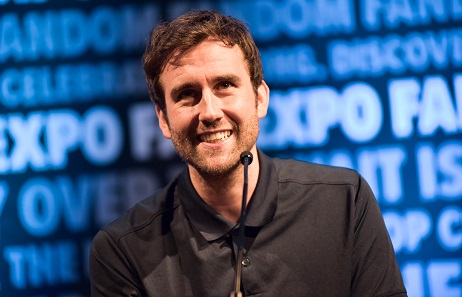 Matthew Lewis sitting in front of a small microphone, smiling and looking up. Wearing a black button down shirt.