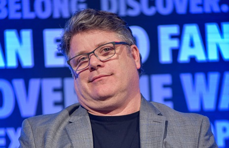 Sean Astin smirking, with a black t-shirt and grey suit jacket on.
