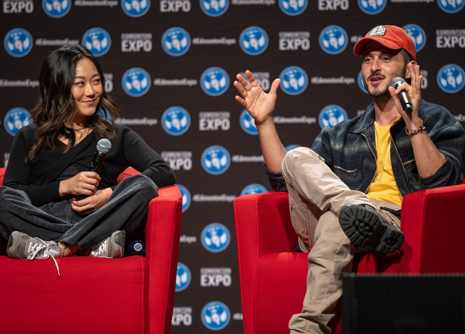 Tomer Capone (wearing a zip up jacket, yellow shirt, khakis and a red baseball cap) is sitting in a red chair talking into a microphone with his hands in a "the fish was this big" motion. Karen Fukuhara (wearing a long sleeve black shirt and some dark comfy jeans) is sitting criss crossed on the red chair and is smiling at Tomer.