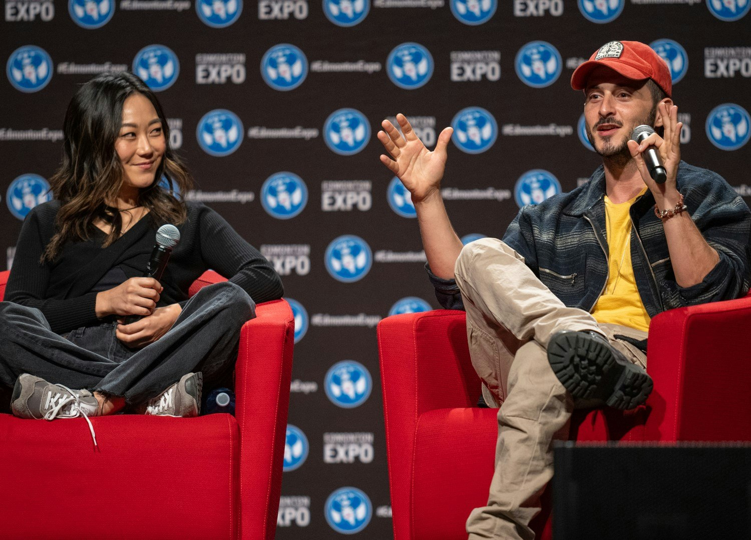 Tomer Capone (wearing a zip up jacket, yellow shirt, khakis and a red baseball cap) is sitting in a red chair talking into a microphone with his hands in a "the fish was this big" motion. Karen Fukuhara (wearing a long sleeve black shirt and some dark comfy jeans) is sitting criss crossed on the red chair and is smiling at Tomer.