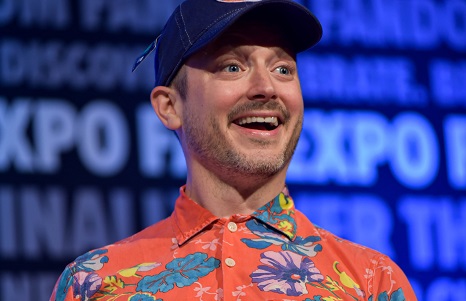 Elijah Wood smiling, with a blue baseball cap and orange, floral, Hawaiian shirt on