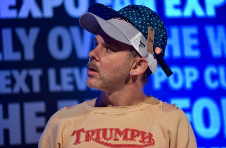 Dominic Monaghan wearing two baseball caps on top of each other, the first grey, and the second one blue with polka dotes. Wearing a beige t-shirt that says "TRIUMPH" in red letters