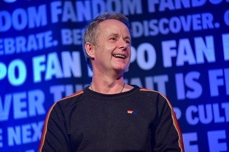 Billy Boyd looking off to the right, smiling and laughing. Wearing a black sweatshirt with rainbow stripes down the middle of the sleeves.