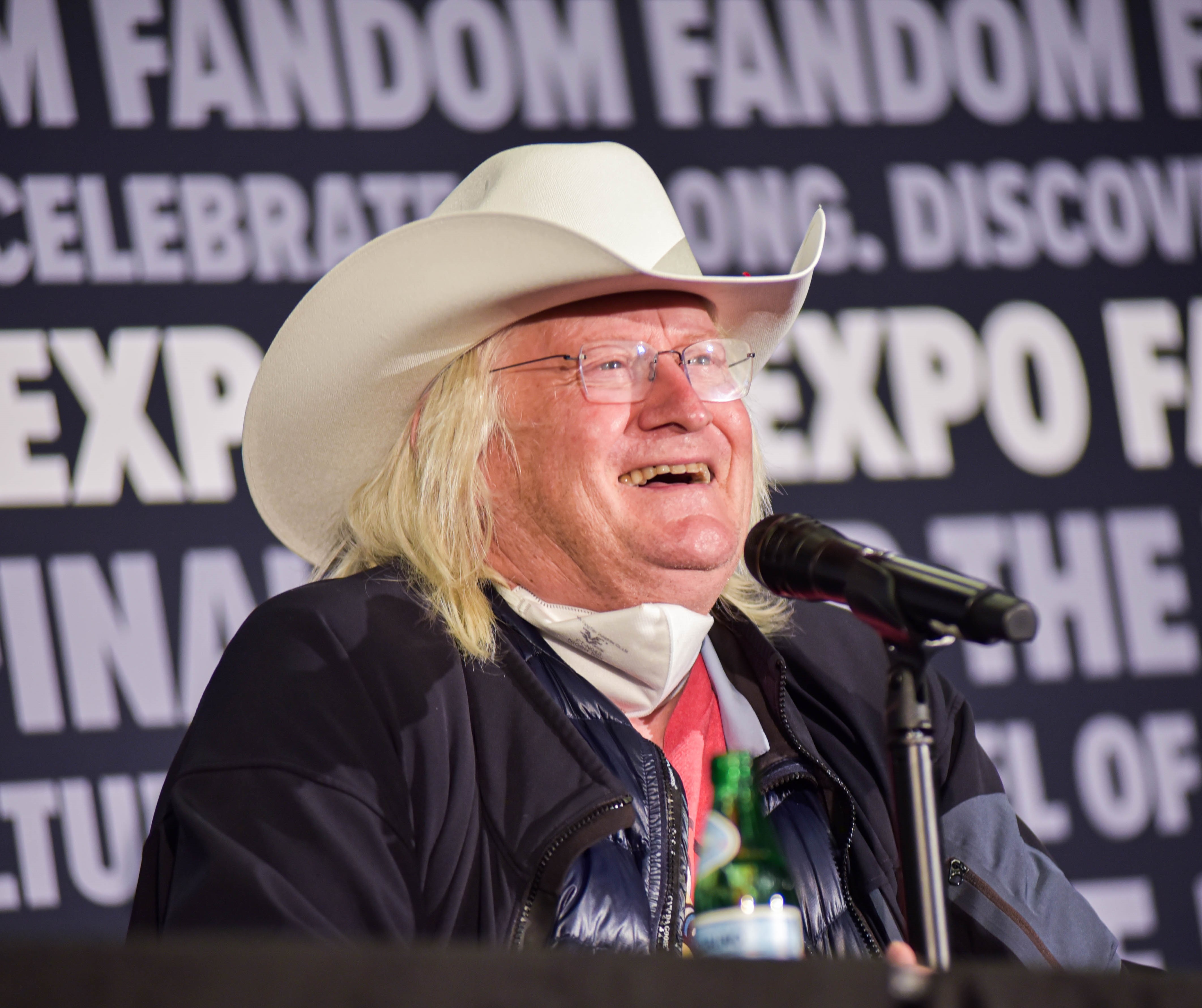 Charles Martinet in a white cowboy hat and a black coat with a microphone in front of him. He is smiling.