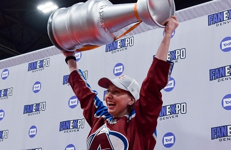 woman standing with a large prop-version of the Stanley Cup above her head, dressed in a Colorado Avalanche Jersey