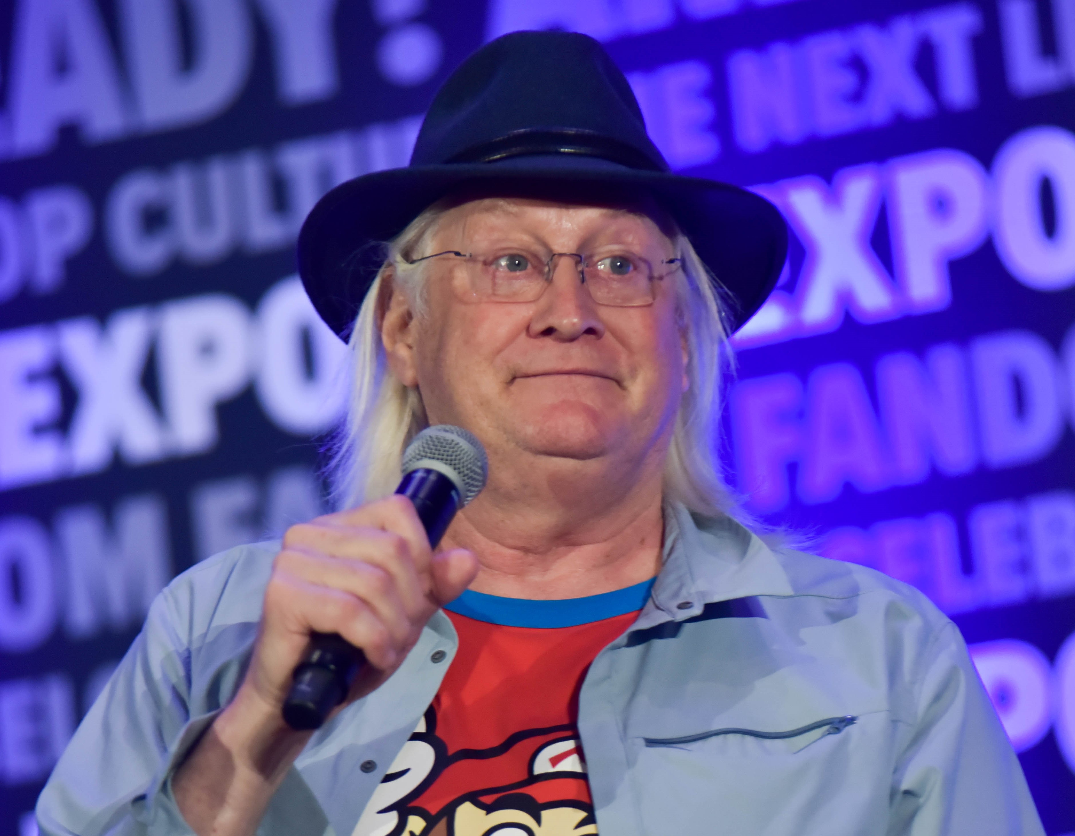 Charles Martinet in a black fedora, blue button up and a red Mario shirt. He is holding a microphone.