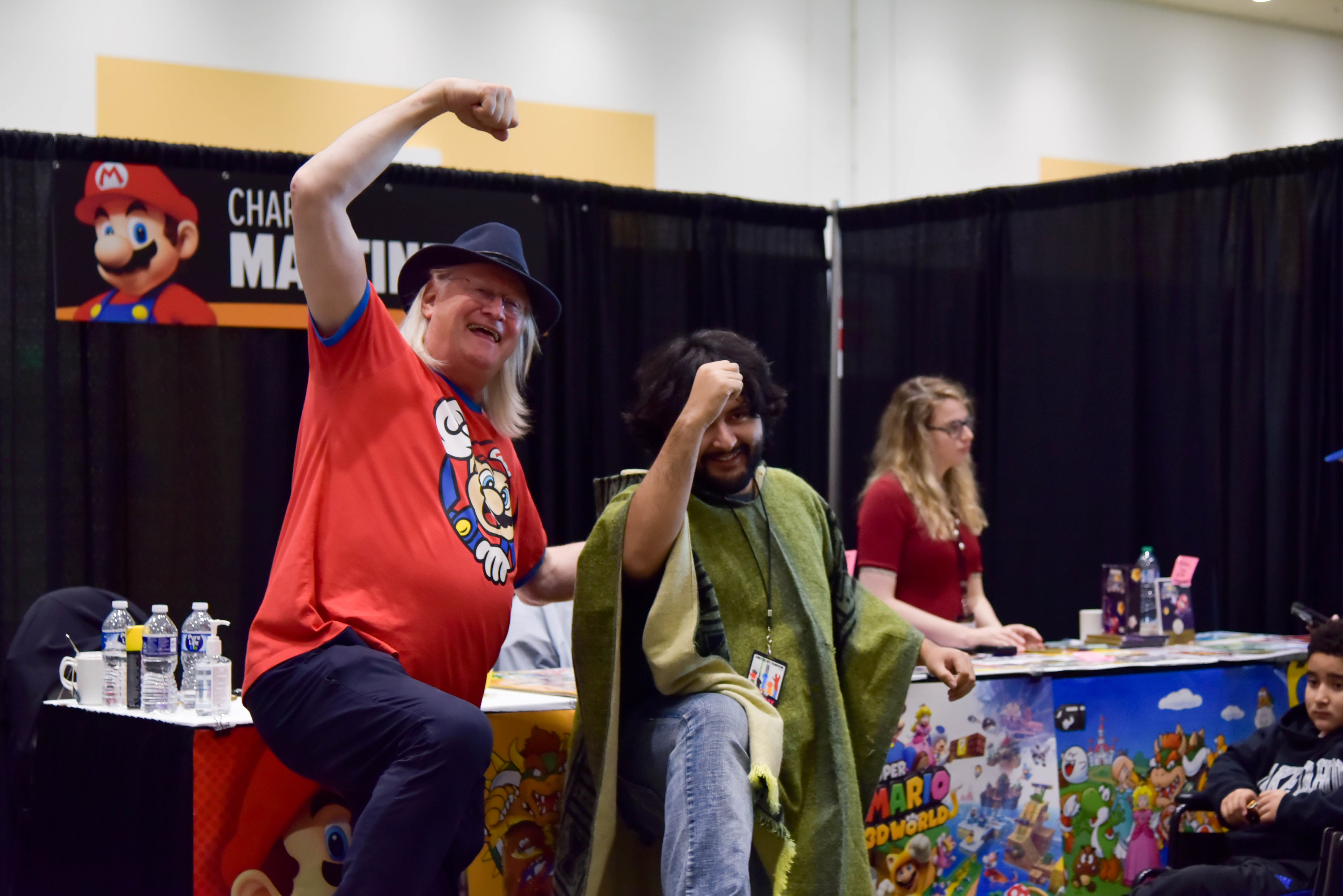 Charles Martinet in a black fedora and a red Mario shirt. He is posing in the Mario jump pose in front of his autograph table with a fan dressed in jeans and a green poncho.