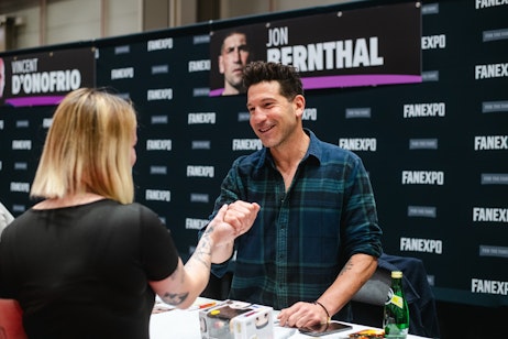 The image shows Jon Bernthal at a fan expo event, interacting with a fan across a signing table. He is smiling warmly and reaching out to the fan in a friendly manner, suggesting a handshake or a gesture of appreciation. Jon is casually dressed in a plaid shirt, and behind him is a backdrop with names, likely indicating the guests of the event. The atmosphere is that of a meet-and-greet area at a convention where fans have the opportunity to meet their favorite personalities. The focus is on the genuine connection being made, highlighting the personal experience such fan events can offer.