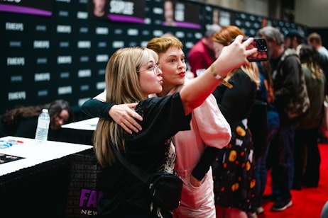 The image captures a fan and actress Rose McGowan taking a selfie together. The woman on the left has long blonde hair, wears glasses, and is dressed in a black top. She is holding the smartphone and embracing Rose on the right. Rose McGowan has short, styled hair, is wearing a pink top with a coral-colored overtop, and red lipstick. They are both puckering their lips for the photo and seem to be enjoying the moment. In the background, there's a banner with the word "FAN" visible, indicating they might be at a fan convention or similar event. Other attendees and possibly event staff can be seen in the background, suggesting a lively, public atmosphere.