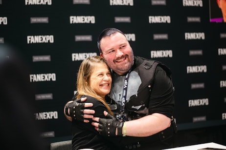 The image shows a heartfelt embrace between Linda Hamilton and a man at an event. Linda is wearing a black top and has a joyful expression on her face as she hugs the man, who is smiling broadly and wearing a black vest with a VIP badge indicating special access at the event. They seem to be sharing a moment of genuine affection or celebration. The backdrop with the "FAN EXPO" logo suggests that this is taking place at a fan convention or similar gathering where attendees can meet and interact closely with guests or celebrities. The warmth and happiness of their interaction is the focal point of the image.