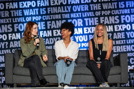 The image captures a panel discussion at a fan expo event, featuring Felicia Day, Sonequa Martin-Green, and Michelle Hurd engaged in a lively conversation. Felicia Day is on the left, speaking into a microphone, likely sharing an anecdote, or answering a question, while Sonequa and Michelle are listening, their faces lit with smiles that suggest enjoyment and engagement in the discussion. Their casual yet stylish attire and relaxed body language, along with the microphones in hand, indicate they are either panelists or special guests at the event. The background features the event's branding, emphasizing the focus on pop culture and the community atmosphere of these conventions. The overall mood is cheerful and animated, with a sense of camaraderie among the speakers.
