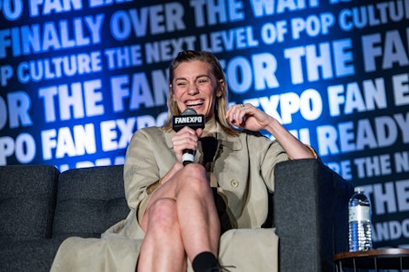 The image features Katee Sackhoff seated, speaking into a microphone with the "FAN EXPO" logo on it, suggesting she is at a fan convention or public interview. She is smiling broadly, mid-laughter, conveying a sense of joy and engagement. Her attire appears to be a stylish, beige trench coat, and her relaxed posture and candid expression suggest she's sharing an amusing story or answering a question with good humor. The backdrop, emblazoned with text related to pop culture and fandom, supports the idea that this is a special event where fans gather to interact with personalities from various entertainment sectors.