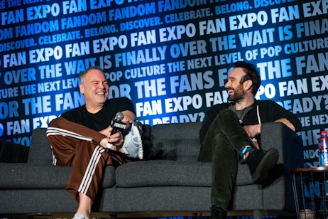 The image features Charlie Cox and Vincent D’Onofrio sharing a joyful moment, seated, and laughing together on stage at a FAN EXPO event. Vincent, on the left, is holding a microphone, dressed casually, and reclining comfortably, while Charlie, on the right, also smiling broadly, appears to be engaging in a light-hearted conversation with him. The backdrop with the text "FAN EXPO" suggests they are part of a panel or a discussion, and their relaxed body language and candid expressions convey a friendly and entertaining atmosphere. The setting is typical of a fan convention where celebrities and industry professionals interact with fans and discuss various aspects of pop culture.