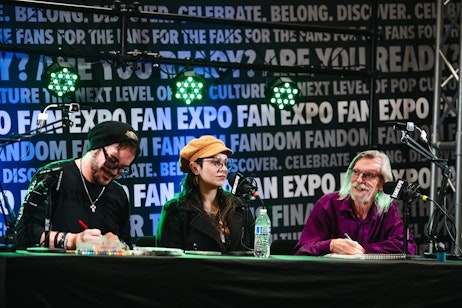The image depicts three individuals seated at a table, likely part of a panel discussion at a fan expo or convention, as indicated by the repeating "FAN EXPO" logo on the backdrop. The person on the left appears to be signing autographs or creating artwork, the middle person is a woman wearing glasses and a mustard-colored beret, and the individual on the right is an older man with long white hair and a beard, smiling as he looks on. The setup suggests they could be artists, writers, or personalities within the pop culture realm, engaging with fans or participating in a panel. The atmosphere is professional, highlighted by stage lighting and microphones in front of each panelist.